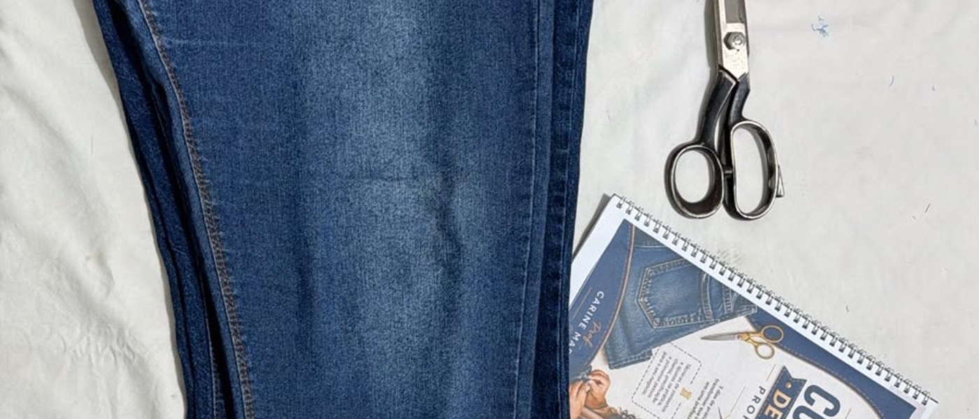 Calça jeans, tesoura de alfaiate, linha e a apostila da Imersão sobre a mesa